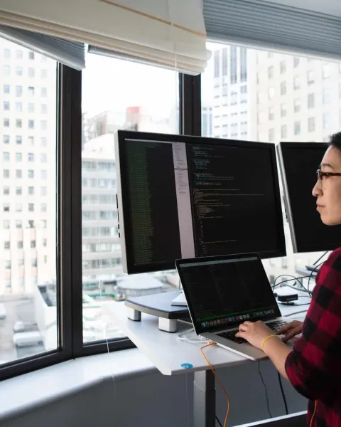Mujer programando en frente de una ventana, utilizando varias pantallas.